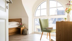 A bedroom with a wooden bed, a green chair, a desk with a vase of flowers, and a large arched window.
