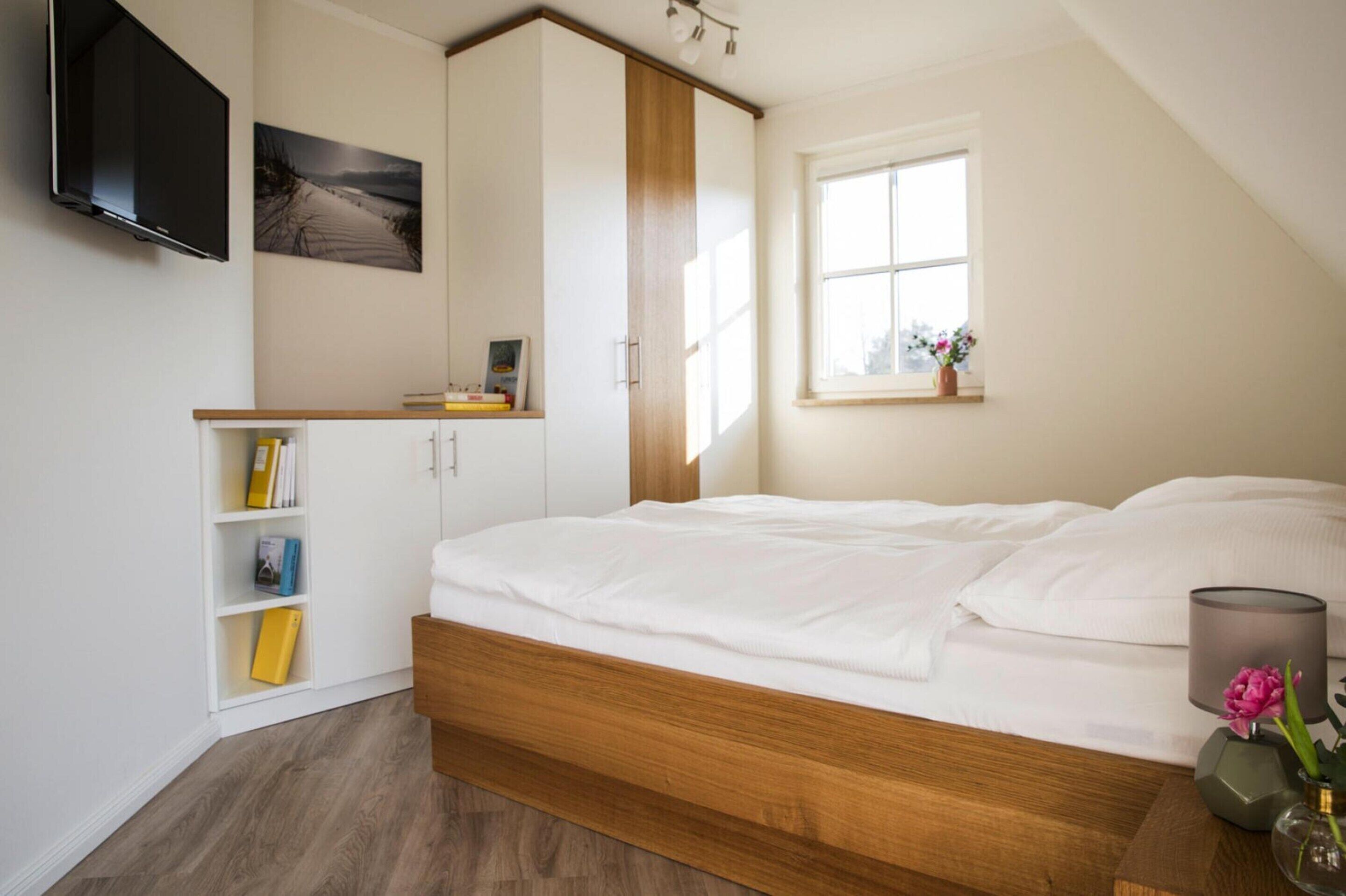 Un dormitorio con cama de madera, paredes blancas, televisor montado en la pared, ventana con vistas y estantería con libros.
