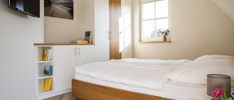 Un dormitorio con cama de madera, paredes blancas, televisor montado en la pared, ventana con vistas y estantería con libros.