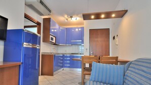 A compact kitchen with blue cabinets, a microwave, and a dining area with a wooden table and chairs. - Apartment in Bibione Near the Beach (San Michele al Tagliamento)