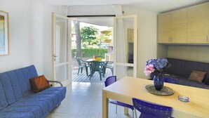 A room with a blue sofa, a wooden dining table with chairs, a vase with flowers, and a view of the outside. - Holiday Village Tivoli in Bibione (San Michele al Tagliamento)