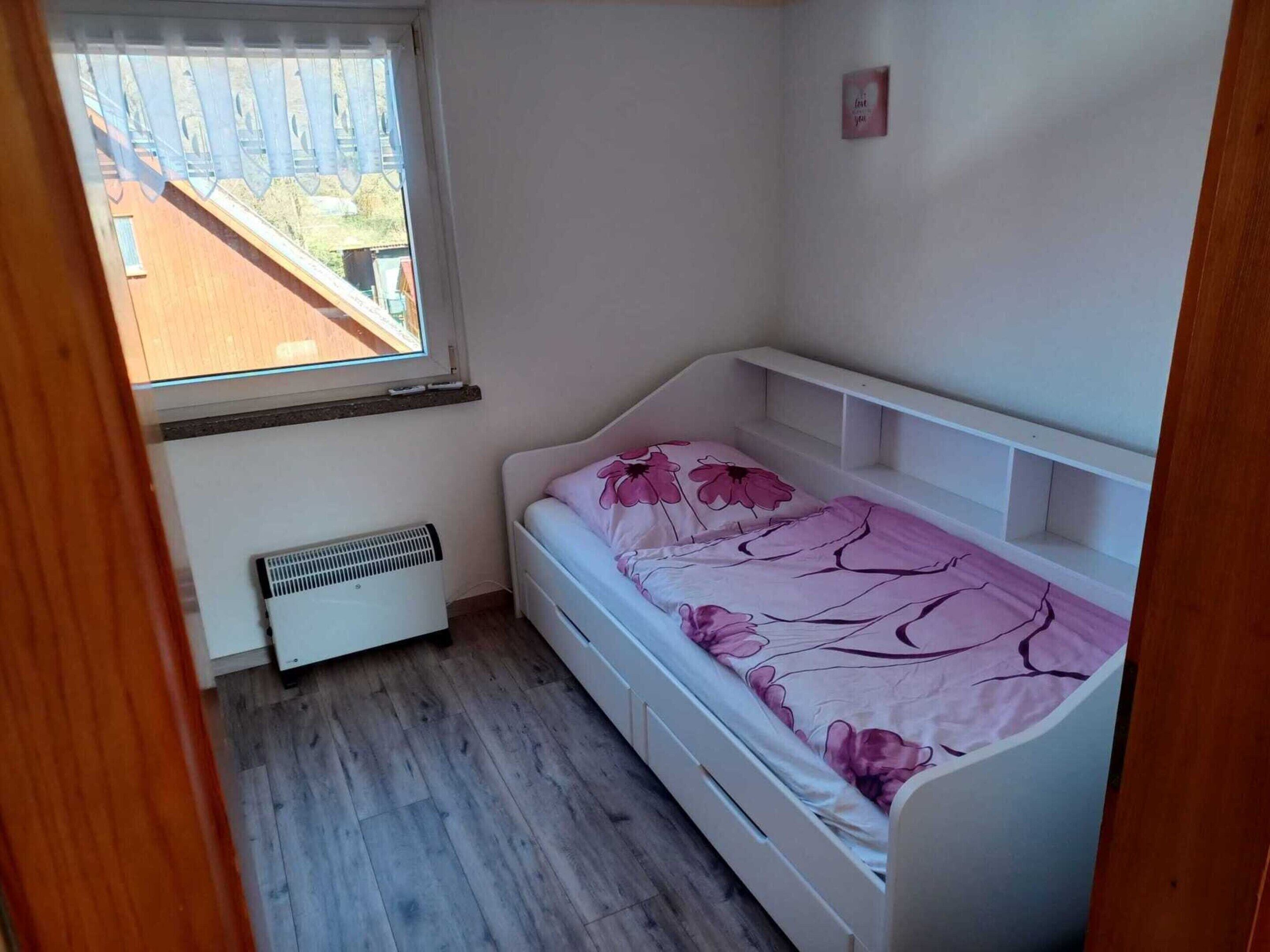 Une petite chambre avec un lit simple, une fenêtre, un radiateur et un mur blanc.