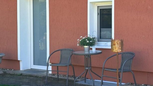 Un espacio al aire libre con dos sillas de metal y una pequeña mesa redonda, una planta en maceta y un accesorio de iluminación decorativo.