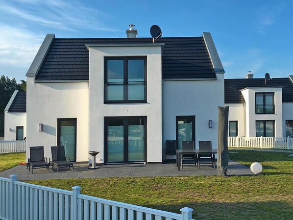 A modern two-story house with a flat roof, large windows, and a patio area with outdoor furniture. - Ferienhaus auf Usedom mit Garten und Hafenblick (Peenemünde)