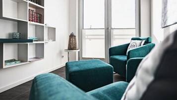 Een moderne woonkamer met teal kleurige banken, een witte boekenkast en een groot raam met zonwering.