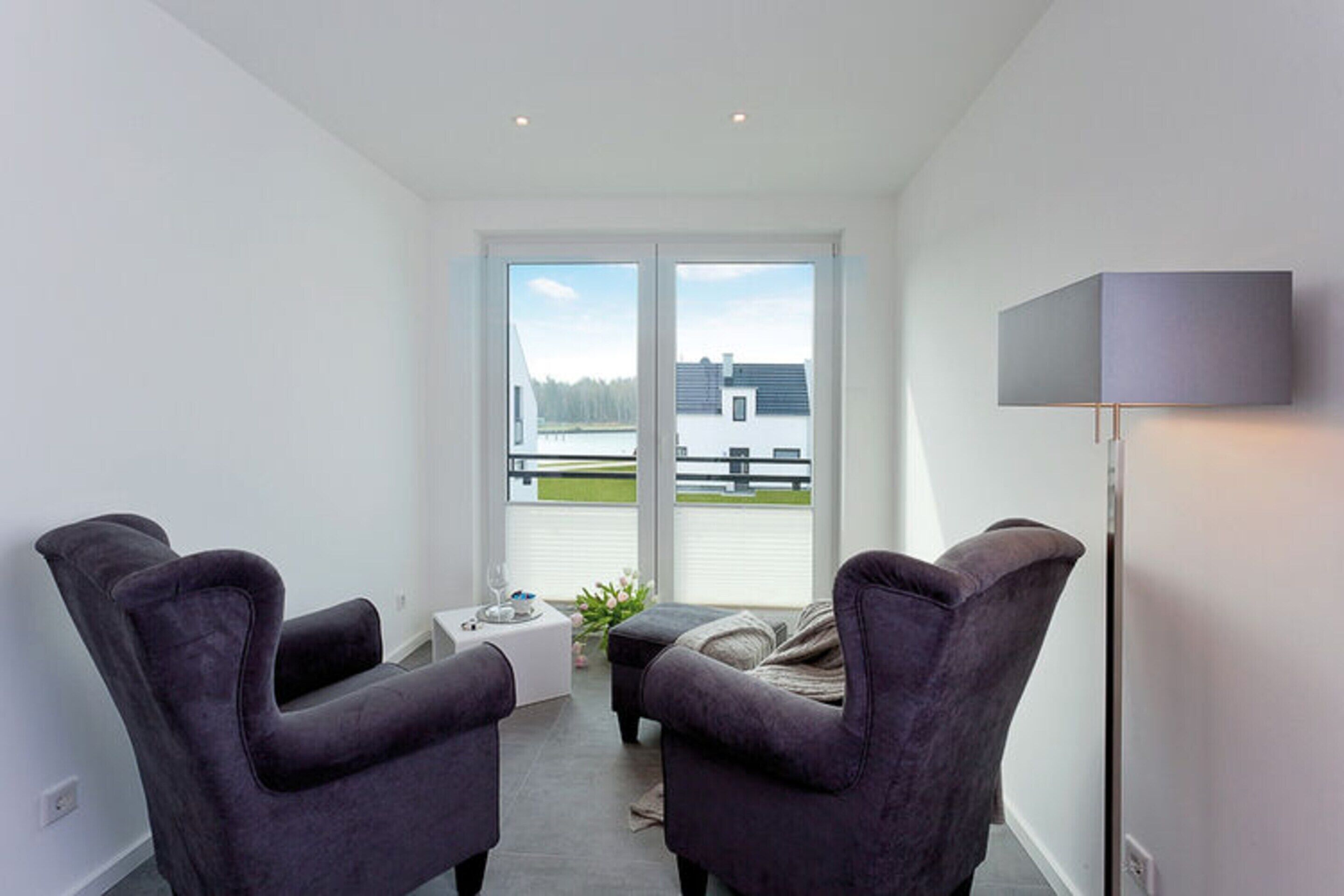 Een moderne kamer met twee donkere fauteuils, een klein tafeltje met een glas, en een vloerlamp. Een groot raam biedt uitzicht op een gebouw en bomen.