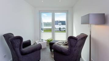 Een moderne kamer met twee donkere fauteuils, een klein tafeltje met een glas, en een vloerlamp. Een groot raam biedt uitzicht op een gebouw en bomen.
