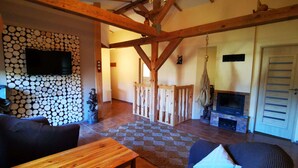 Uma sala de estar rústica com vigas de madeira aparentes, uma parede de pedra com lenha empilhada, uma lareira e uma televisão fixada na parede.