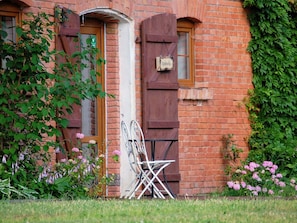 A brick building with a wooden door and shuttered windows, a metal chair, and pink flowers in the foreground. - Holiday Home in the Countryside, Dzierzecin (Postomino)