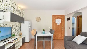 A modern living room with a flat-screen TV, a grey sofa, a dining table with two chairs, and a kitchen area with a fridge and cabinets. - Pretty Apartment in Maspalomas Near the Beach-formerly TUI Ferienhaus (San Bartolomé de Tirajana)