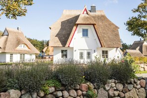 Une maison traditionnelle à toit de chaume, dotée d’une façade blanche, entourée d’un mur de pierre et d’une végétation dense.