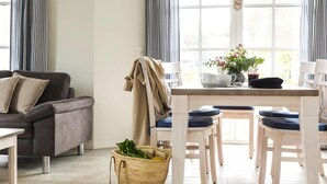 Una sala de estar con un sofá, una mesa de comedor con sillas, una cesta de frutas y un ventanal con cortinas.