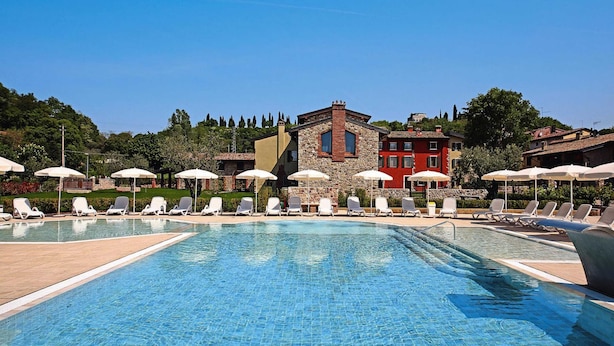 A swimming pool with lounge chairs and umbrellas, surrounded by buildings with stone and red facades. - Apartment in Lazise With Communal Pool (Lazise)