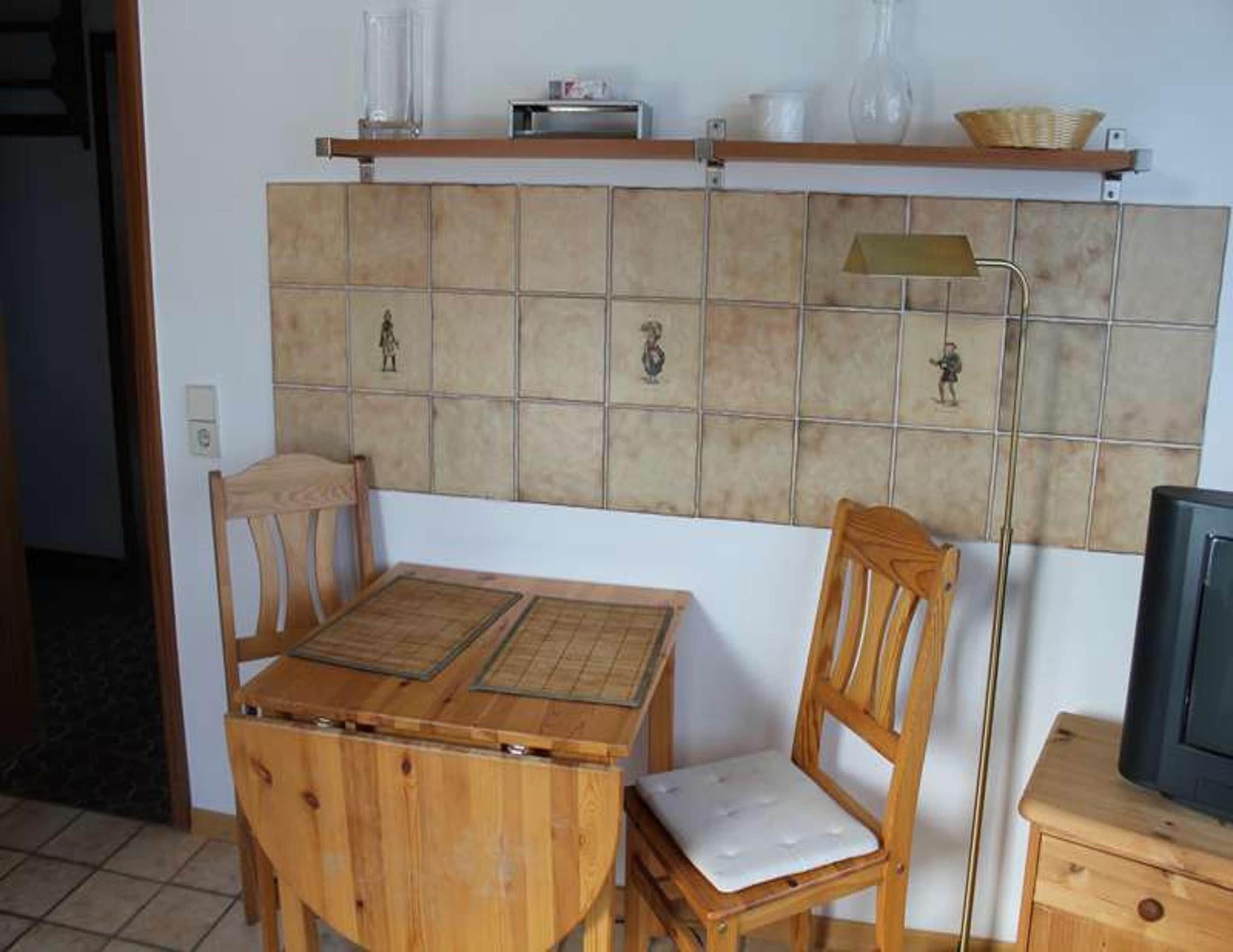 Een kleine keuken met een betegelde spatwand, houten stoelen, een tafel, een vloerlamp en een televisie.