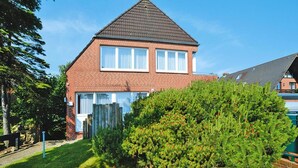 A two-story brick house with a gabled roof, a visible number '6' on the side, and a well-maintained lawn with a large coniferous tree in the foreground. - Ferienwohnung in Westerland (Sylt)