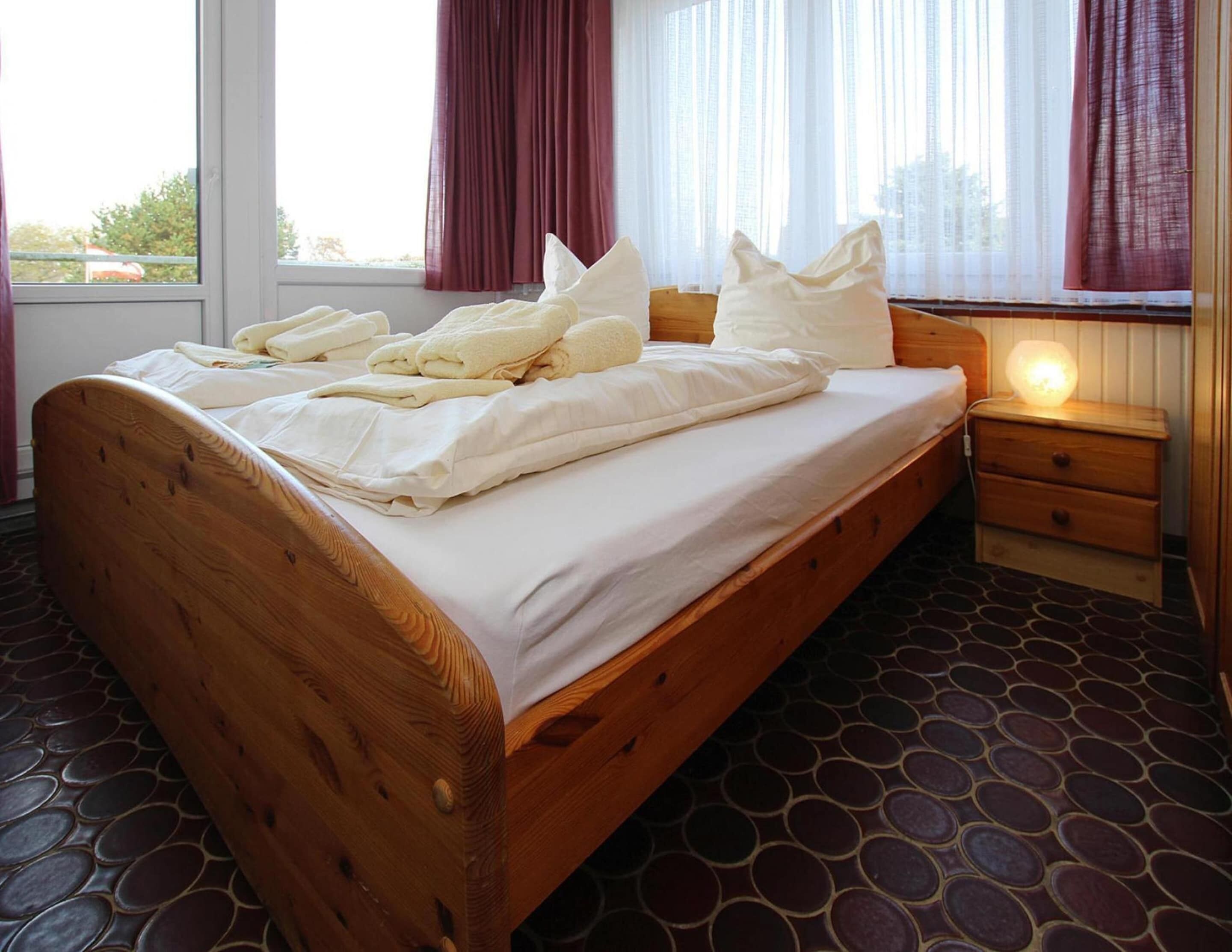 Un lit en bois avec du linge de lit blanc et des oreillers, une table de chevet avec une lampe, et une fenêtre avec des rideaux.