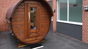 A wooden barrel-shaped structure with a door and a small window, placed on a paved surface with a brick wall and a window in the background. - Ferienwohnung in Westerland (Sylt)