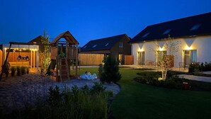 A residential property with a wooden play structure, a covered seating area, and a well-lit patio with outdoor furniture. - Holiday Home Grzybowo Near Beach & Bike Path (Kolobrzeg)