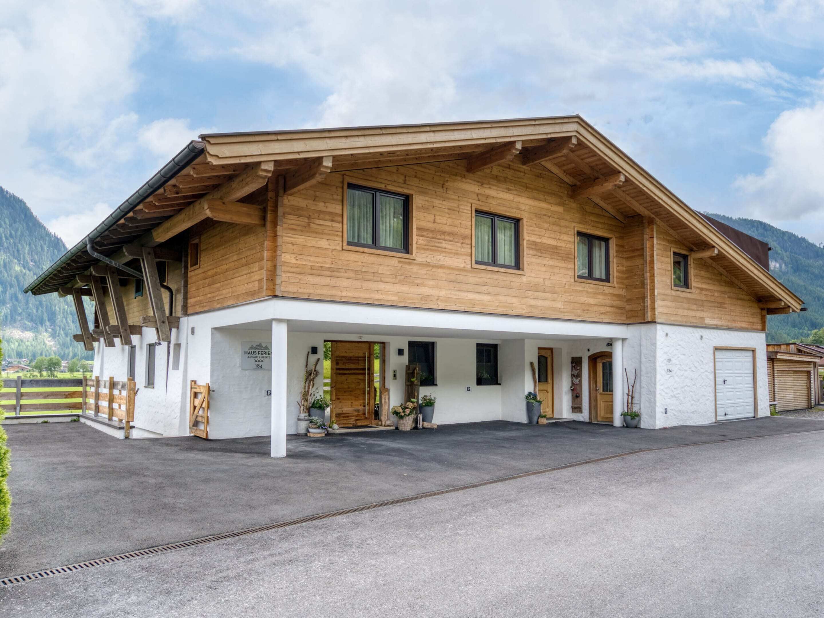 Una casa de madera de dos pisos con un porche cubierto, un garaje y montañas al fondo.