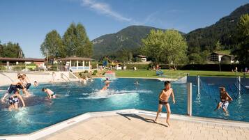 A swimming pool area with multiple people swimming and playing, surrounded by a grassy area and buildings in the background.