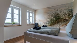 Ein Schlafzimmer mit einem Bett, zwei Fenstern, einer Pflanze und einem Wandbild mit Strandmotiv.