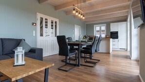 Ein modernes Wohnzimmer mit einem Holzesstisch, schwarzen Stühlen und einem grauen Sofa. Es gibt eine Küchenzeile mit weißen Schränken und einem an der Wand befestigten Fernseher.
