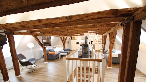 Un loft à aire ouverte avec des poutres en bois apparentes, une rambarde centrale en bois et une petite cuisine située au fond de la pièce.