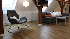 Un appartement moderne de style « loft » avec des poutres apparentes en bois, un fauteuil à bascule noir, un canapé gris avec des coussins orange et un téléviseur à écran plat fixé au plafond.