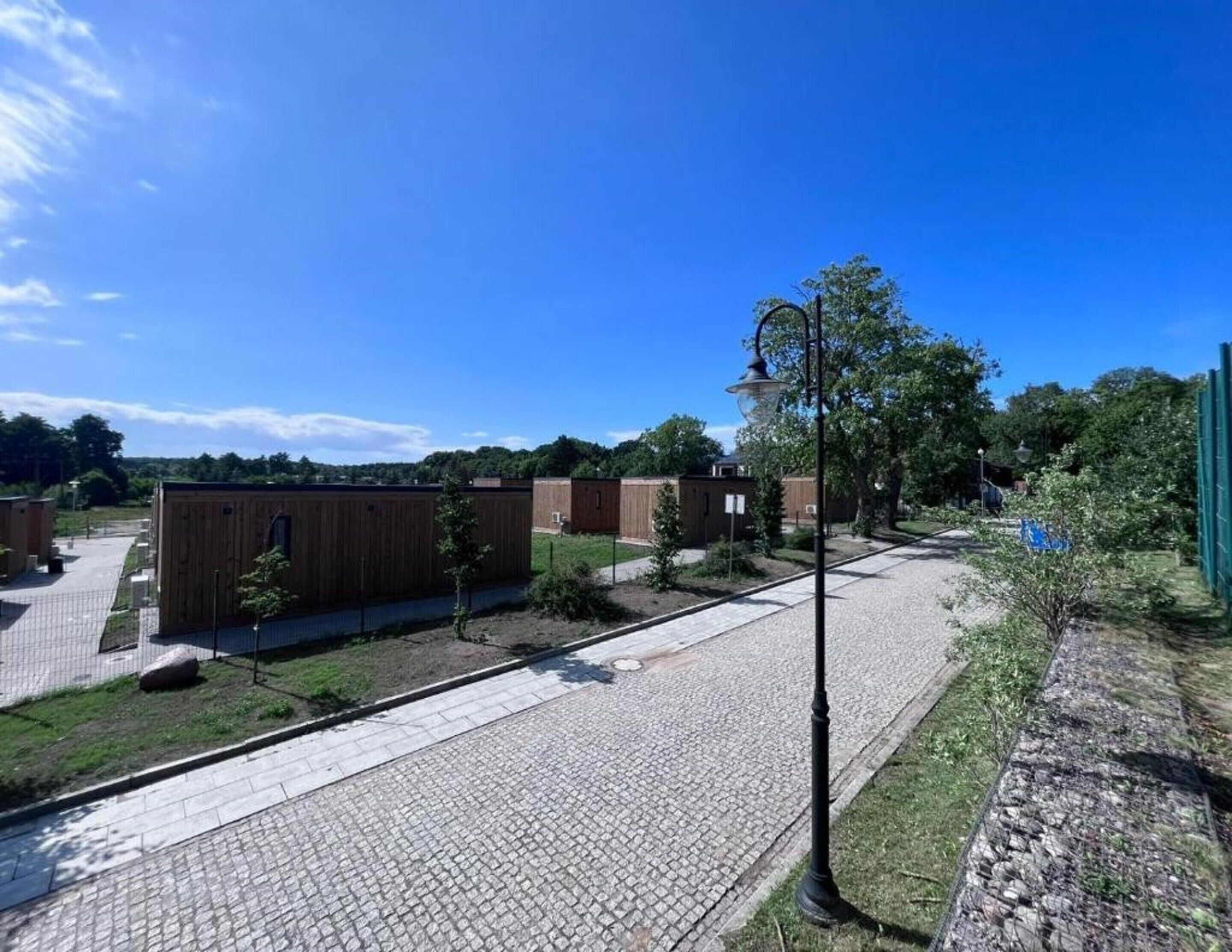 A paved walkway with wooden buildings, a lamp post, and a fenced area with trees.
