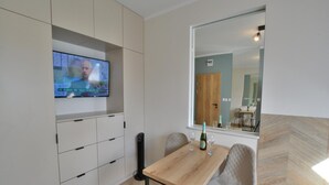 A modern room with a TV mounted on the wall, a wooden table with two chairs, a bottle of champagne, and a large mirror reflecting the room. - Comfortable, 2-person Holiday Studio (Ustronie Morskie)