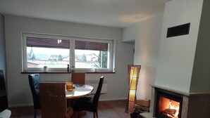 Ein Essbereich mit einem runden Tisch, Korbstühlen und Blick durch ein Fenster. Rechts ist ein Kamin mit Holzscheiten zu sehen.