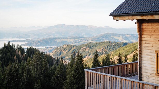 Una cabaña de madera con balcón que da a un valle boscoso y montañas distantes.