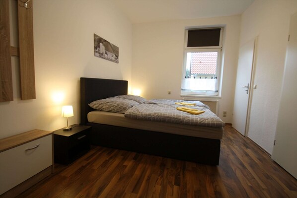 A bedroom with a bed, nightstand, and a window with a view of buildings. - Ferienwohnungen, Malchow (Malchow)