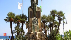 A monument with inscriptions, surrounded by palm trees and flags, located near a body of water. - Apartment in Pietra Ligure With Garage (Pietra Ligure)