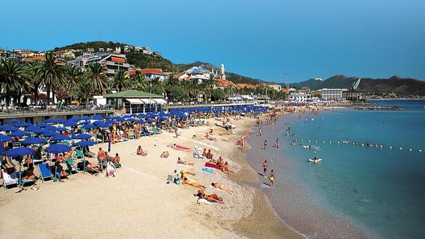 A beach with rows of blue umbrellas, people sunbathing, and a building with a red roof in the background. - Apartment in Pietra Ligure With Garage (Pietra Ligure)