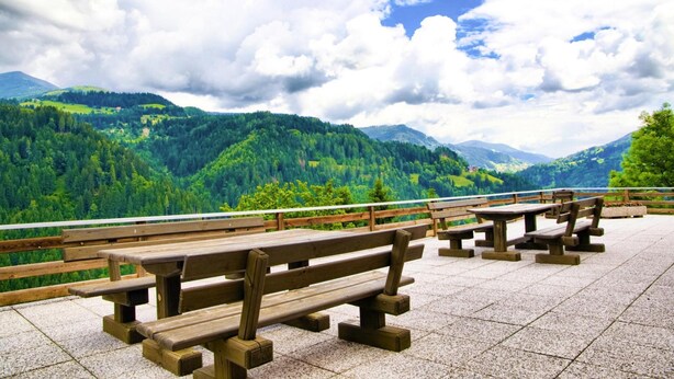 A scenic outdoor seating area with wooden benches and tables, overlooking a mountainous landscape with dense forests and a clear sky. - Farmhouse Near Afritzer See & Nockberge Trails (Treffen)