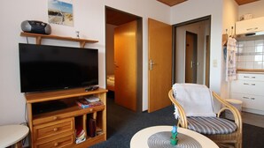 Ett hotellrum med en tv på ett träställ, ett litet bord med en växt, en flätad stol och ett litet kök i bakgrunden.