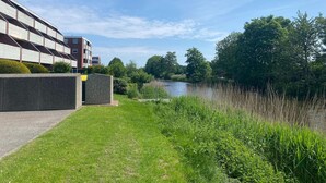 En strandpromenad längs floden med en modern byggnad, frodig grönska och en klar himmel.