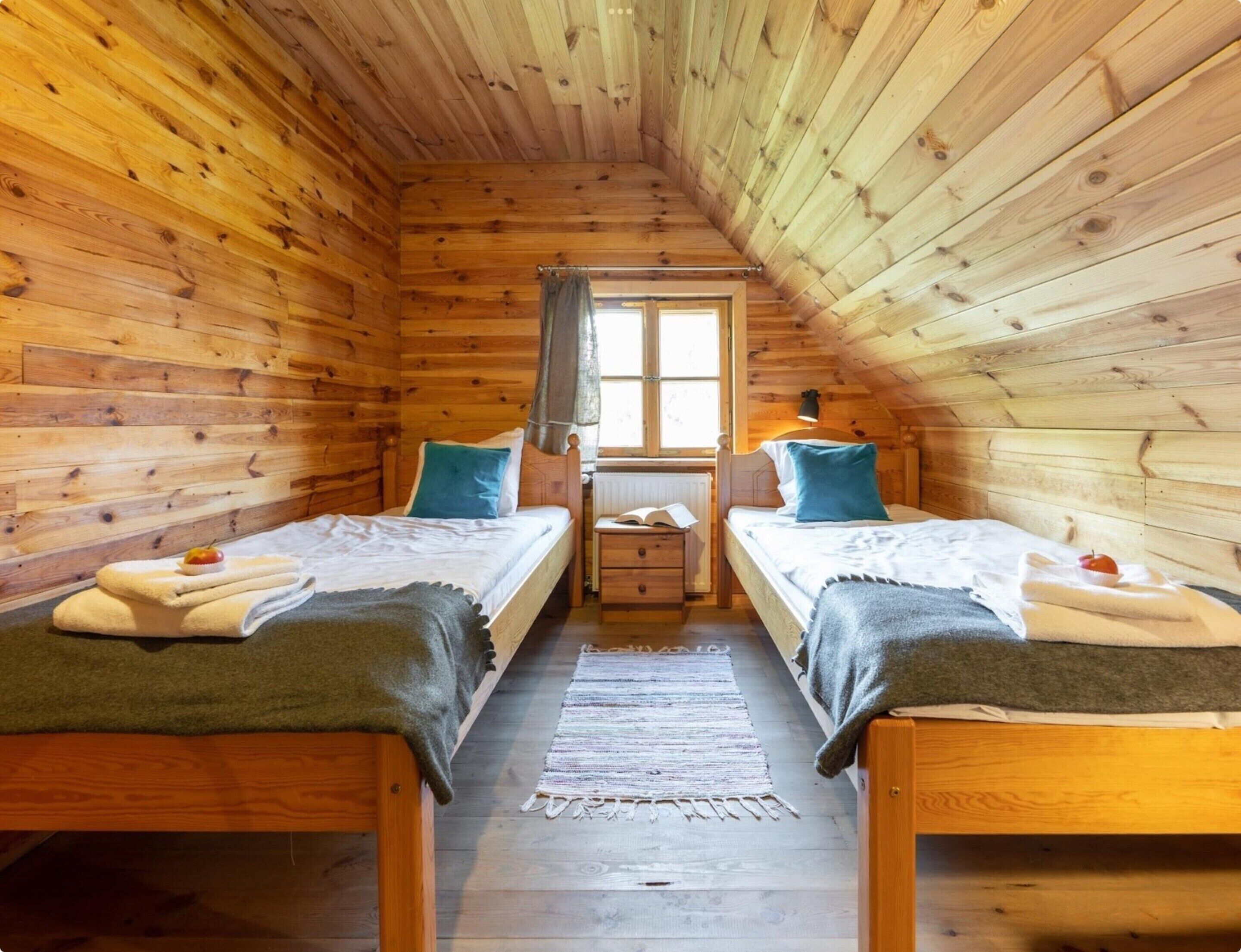 A cozy attic bedroom with two wooden beds, a window with curtains, and a small bedside table with a book.