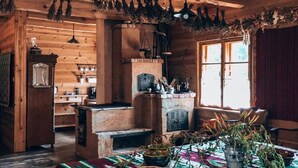Interior rústico de una cabaña de madera con chimenea central, vigas de madera y un comedor con una mesa dispuesta para dos.
