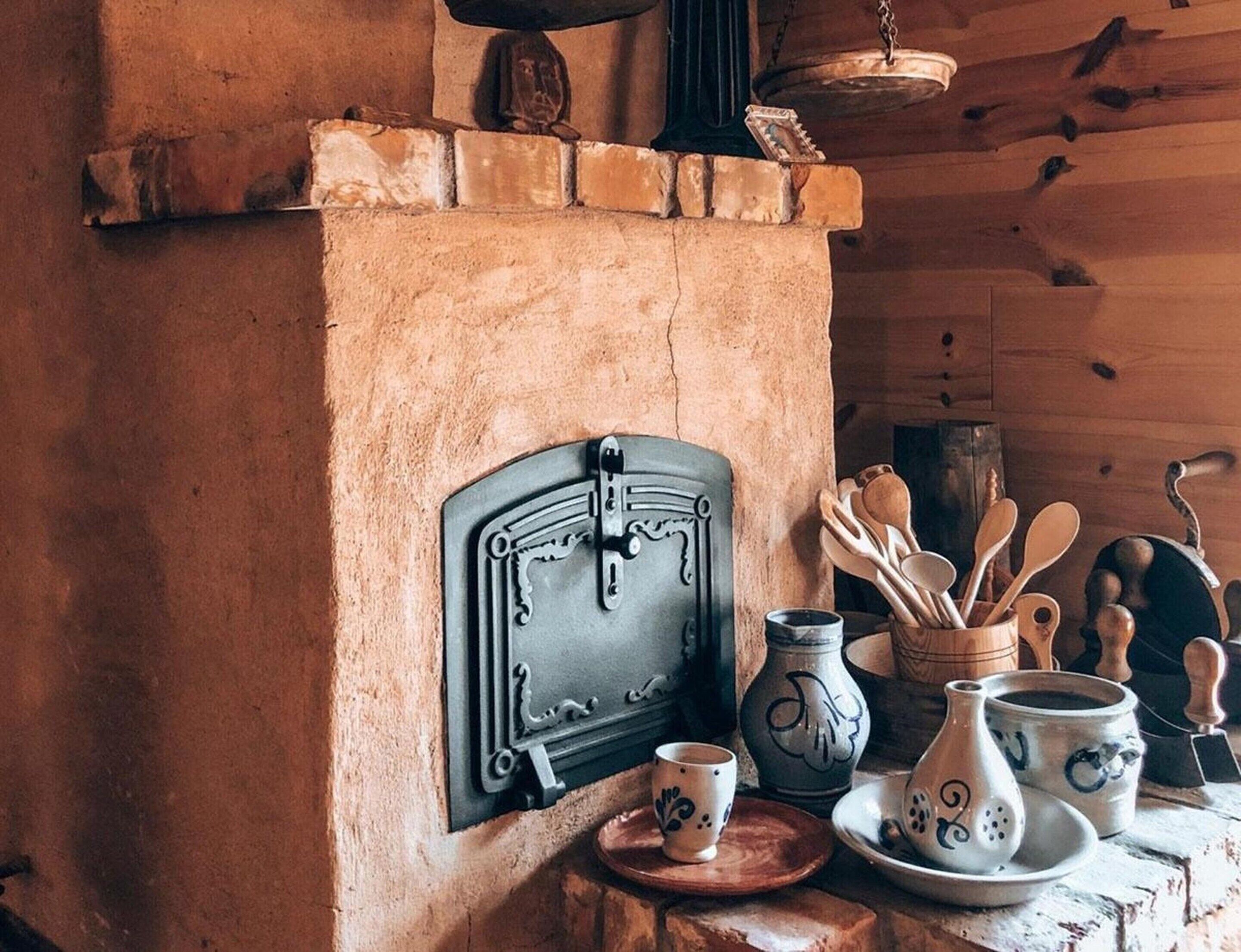 A rustic kitchen with a brick fireplace, a hanging pot rack, and various kitchen utensils and pottery.