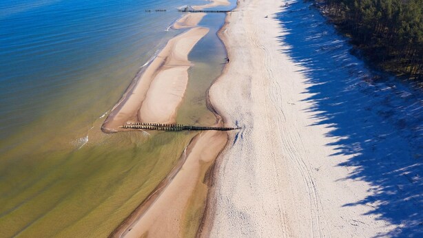 Luftaufnahme eines Strandes mit klarem Wasser, einer Holzsteganlage und einem bewaldeten Ufergebiet.
