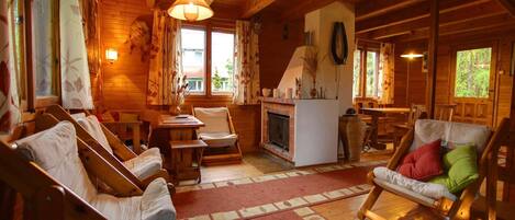 Ein gemütliches Blockhaus-Interieur mit Holzwänden, einem Kamin, einem Essbereich und einer Sitzecke mit Sesseln und einer Couch.