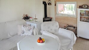 A cozy living room with a white sofa, a covered armchair, a round table with fruit, and a window with a view of greenery. - Holiday Home on Lake Ostrowo Near Baltic Sea (gmina Wolin)
