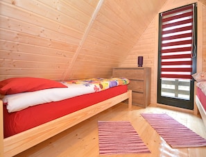 A cozy attic bedroom with a wooden sloped ceiling, a single bed with red bedding, a small wooden dresser, and a window with red blinds. - Holiday Home Near Dabki Beach (Darlowo)