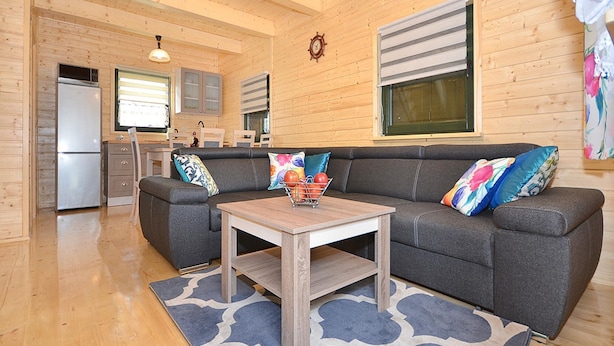 A cozy cabin interior with a grey sofa, wooden ceiling, and a kitchen area with a refrigerator and cabinets. - Holiday Home Near Dabki Beach (Darlowo)