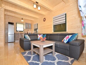Ein gemütliches Blockhaus-Interieur mit einer grauen Couch, Holzbalkendecke und einer Küchenzeile mit Kühlschrank und Schränken.