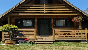 A wooden cabin with a front porch, a black door, and a small garden with flowers and a tree. - Holiday Home Near Dabki Beach (Darlowo)