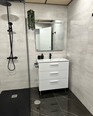Features a sleek vanity with a large mirror, black showerhead, and a potted plant.