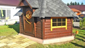 A small wooden cabin with a sloped roof, a wooden door with a circular emblem, and a window with a yellow frame. - Large Holiday Villa With Summer Pool and Sauna (gmina Sianów)